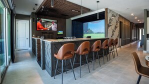 A modern bar area with a long counter, multiple stools, and a television screen displaying a golf course. - Luxury Golf Estate with Simulator, Bowling, Sauna (Reunion)
