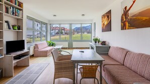 A modern living room with a sofa, a TV, a bookshelf, a dining table, and large windows offering a view of the outdoors.
