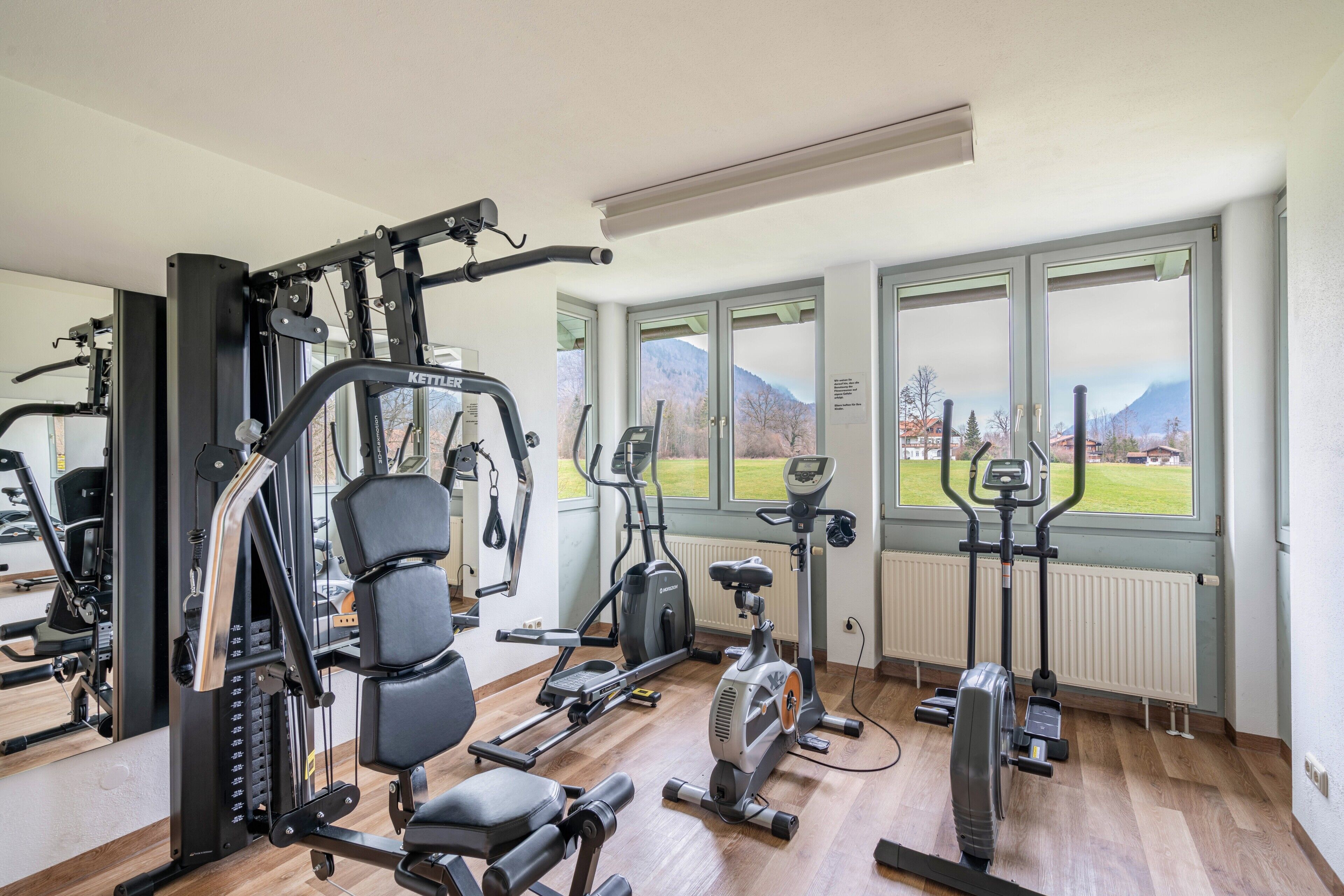 A gym with various exercise equipment, large windows with a view of mountains, and a wooden floor.