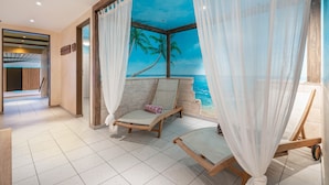 A room with a beach mural, two lounge chairs, a wooden cart, and a view of the ocean.