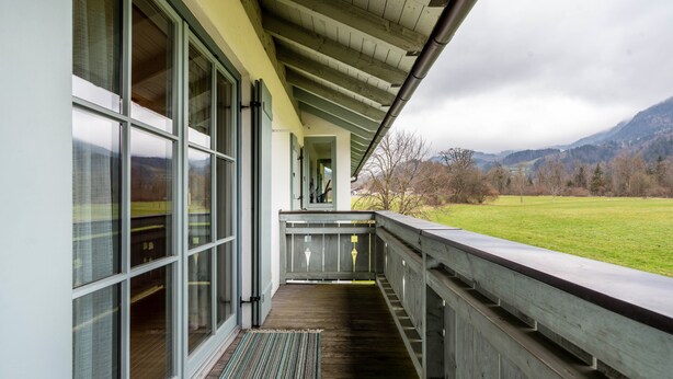 A covered porch with large windows, a wooden deck, and a view of a grassy field and mountains.