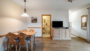 Uma sala de jantar com uma mesa e cadeiras de madeira, uma televisão sobre um móvel, e um banheiro visível através de uma porta aberta.