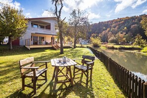 A lakeside property with a wooden deck, a table with chairs, and a building with large windows.
