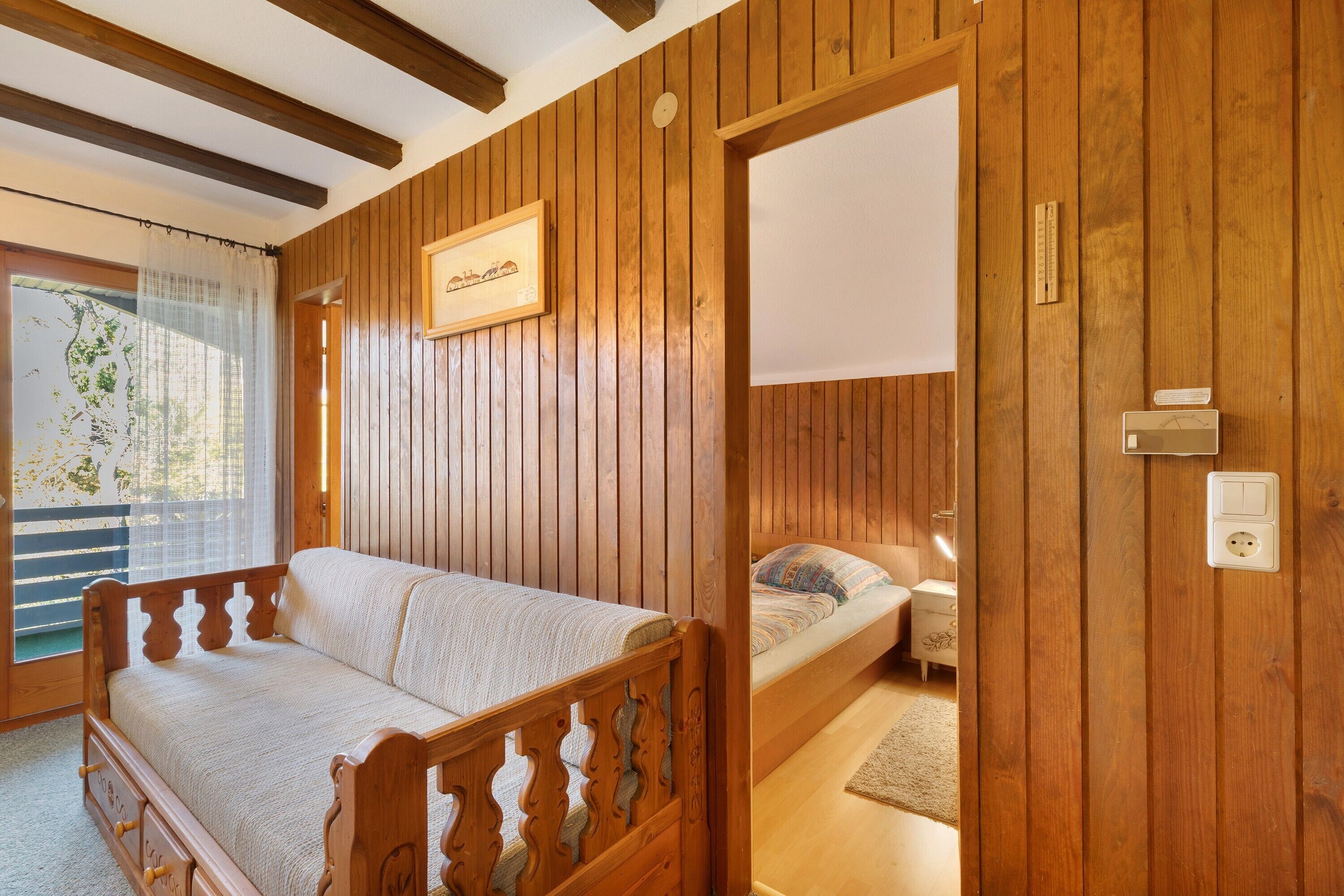 Ein Zimmer mit Holzwänden, einem Bett, einer Couch, einem Fenster mit Vorhängen und einem an der Wand befestigten Thermostat.