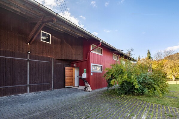 A red building with a wooden door and windows, a paved driveway, and a tree in the foreground.