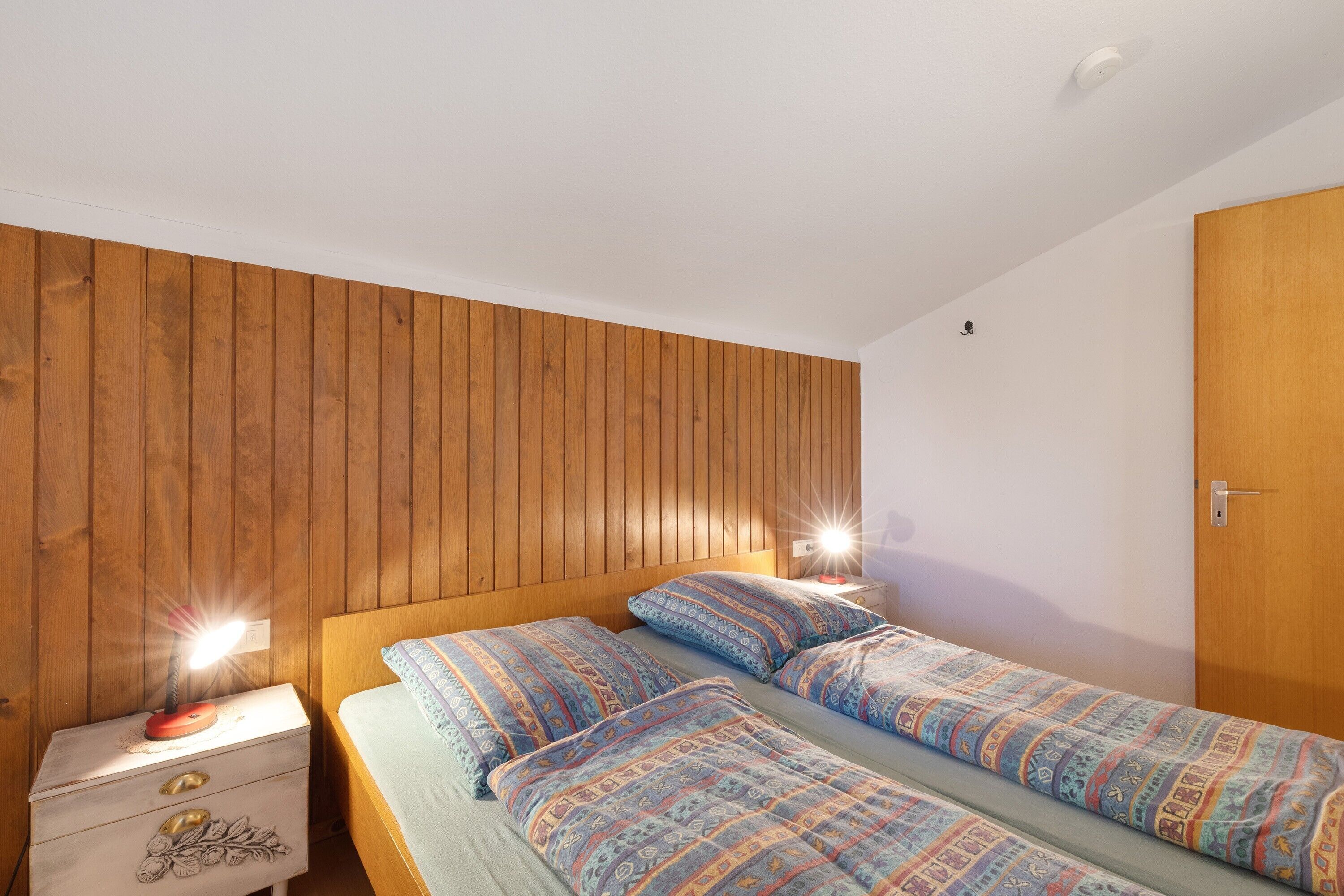 Une chambre avec deux lits, un lambris en bois sur le mur, une table de chevet avec une lampe et une porte.