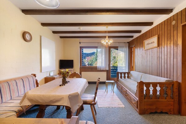 Una habitación con un banco de madera, una mesa con mantel, un televisor y una ventana con cortinas.