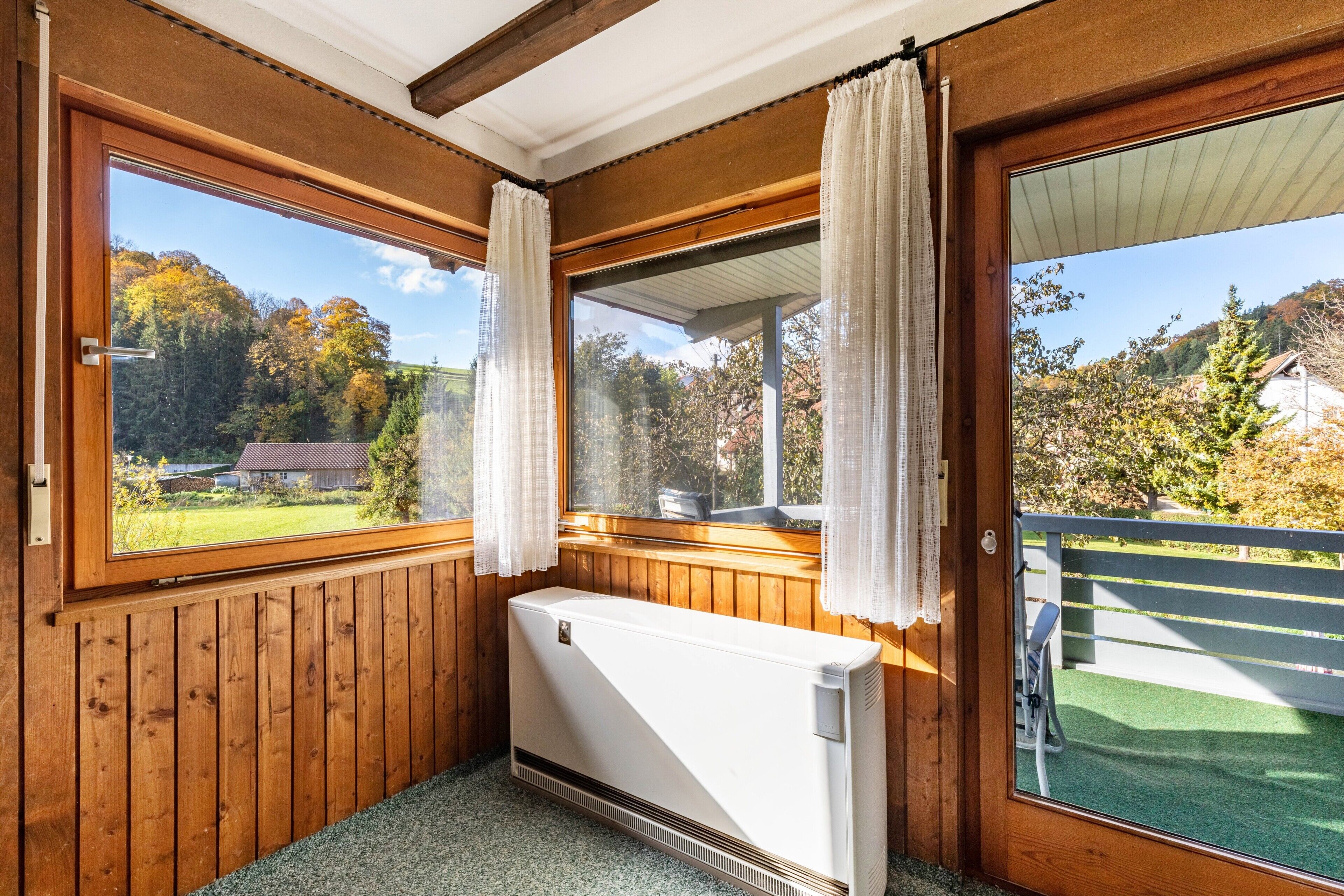 Ein Zimmer mit Holzwänden, einem Heizkörper, einem Fenster mit Vorhängen und Blick auf Bäume und ein Haus.