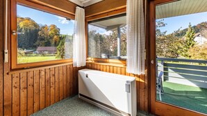 A room with wooden walls, a radiator, a window with curtains, and a view of trees and a house. - Apartment '3' with Shared Garden and Wi-Fi (Hayingen-Indelhausen)