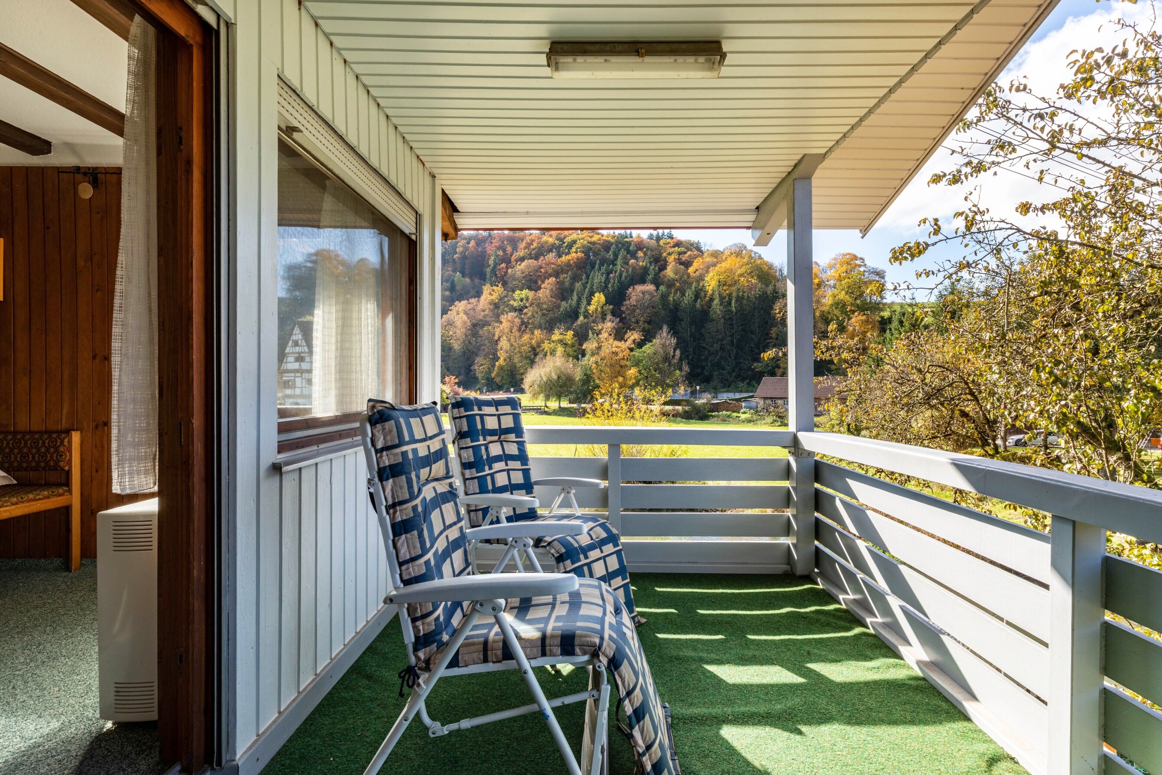Un balcon abrité, doté de deux chaises, d’une balustrade blanche et offrant une vue sur une colline boisée.
