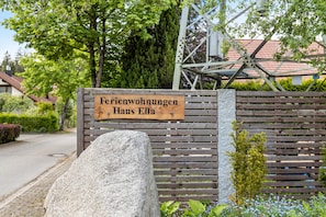 A wooden sign with the text 'Ferienwohnungen Haus Ella' in front of a wooden fence with a stone base.