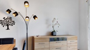 Une pièce comprenant une table à manger en bois, une chaise bleue, un meuble en bois avec des tiroirs, un lampadaire et des éléments de décoration murale, notamment un arbre et des orchidées.