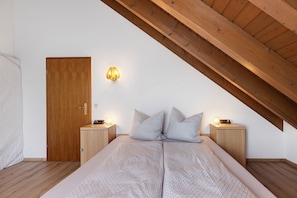 A bedroom with a sloped ceiling, a wooden door, a bedside table with a lamp, a bed with a patterned bedspread, and two pillows.