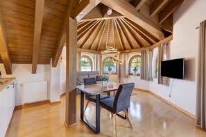 Een ruime kamer met een houten plafond, een eettafel met stoelen, een bank, een aan de muur bevestigde televisie en grote ramen.