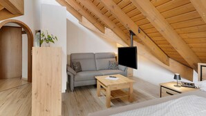 Un dormitorio tipo loft con techo de madera, un sofá gris, una mesita de centro de madera, una mesita de noche con lámpara y una cama con colcha blanca.