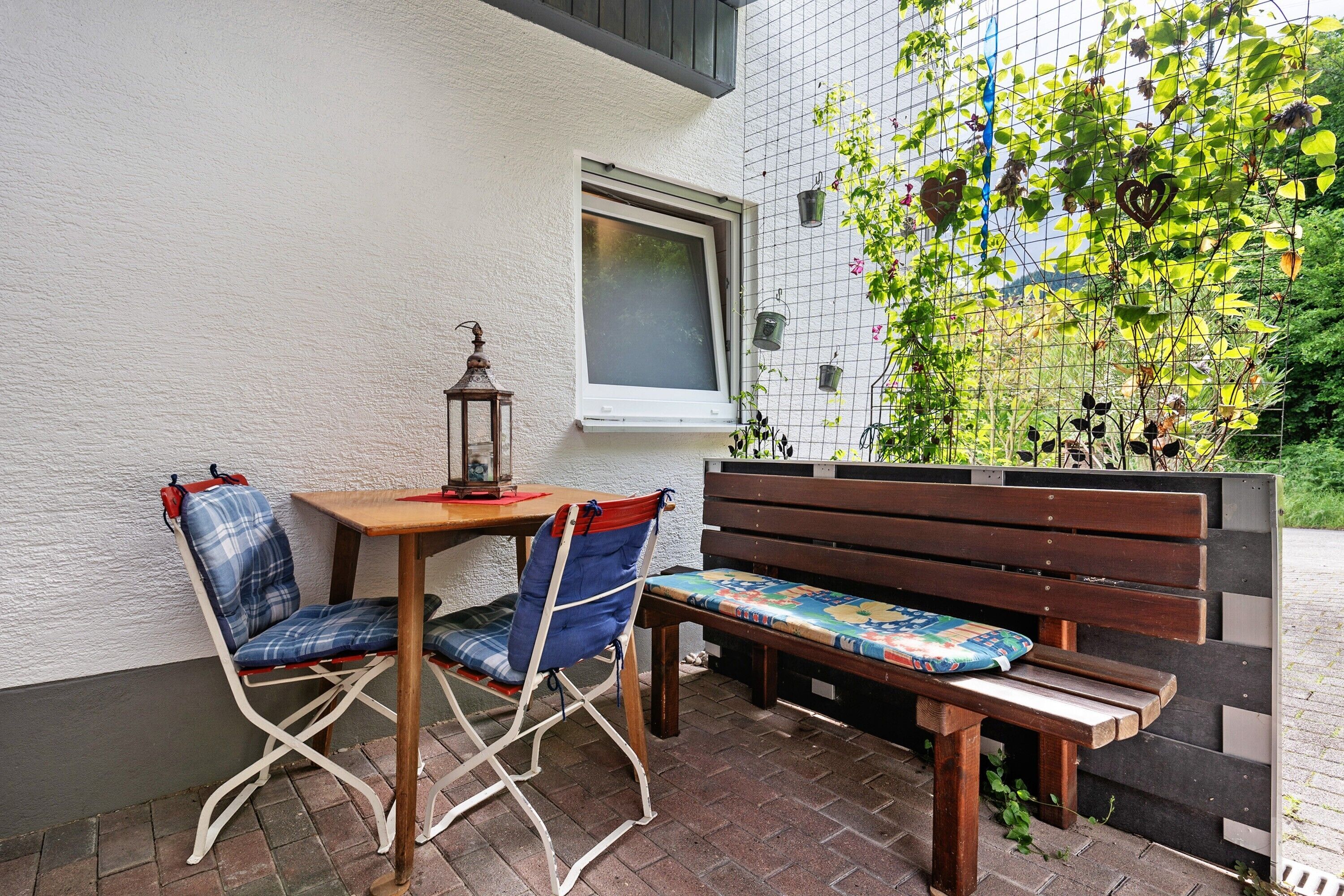 Une terrasse avec une table en bois et deux chaises, un banc, une lanterne et une fenêtre donnant sur un paysage verdoyant.