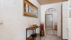A hallway with a textured wall, a framed bulletin board, a wooden console table with decorative items, a vase with plants, and a view into a room with a chair and a table. - Apartment 'Zapf' with Shared Terrace, Shared Garden and Wi-Fi (Forbach)