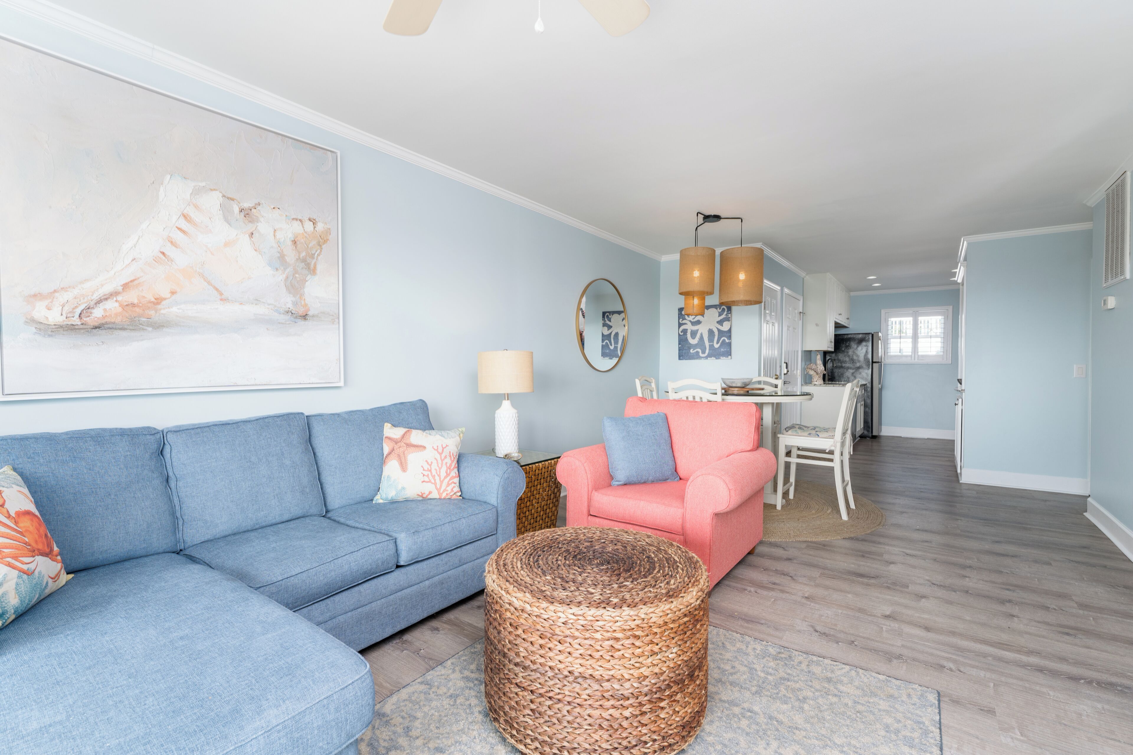 Ein Wohnzimmer mit einem blauen Sofa, einem rosa Sessel und einem gewebten Hocker. Im Hintergrund ist ein Essbereich mit Tisch und Stühlen zu sehen.