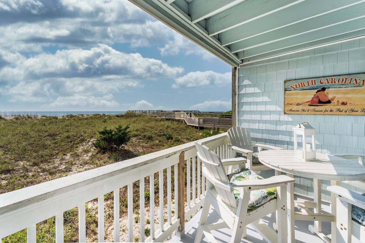 A beachside balcony with a view of the beach and ocean, featuring a wooden deck, white chairs, and a table with a lamp.