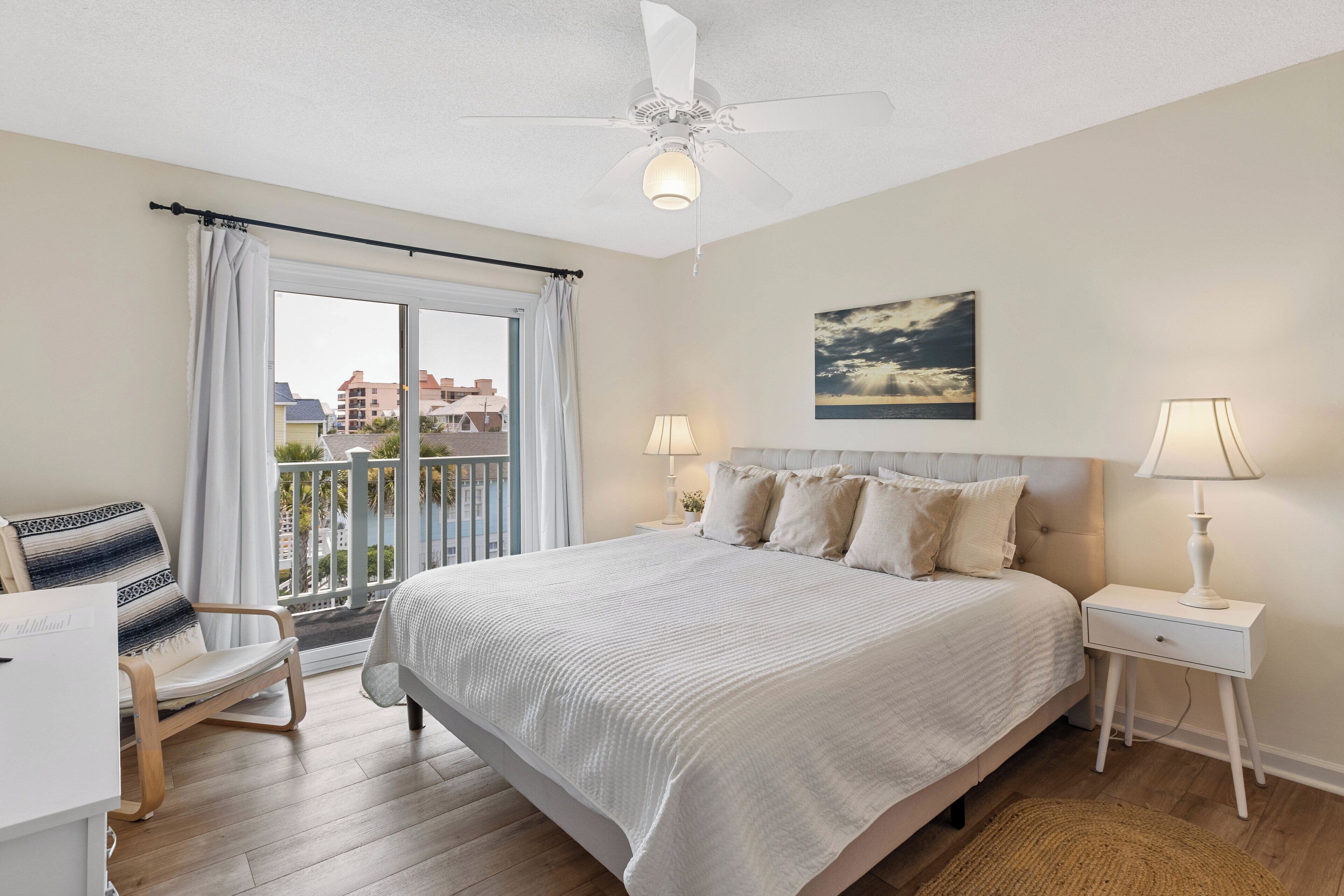 A bedroom with a bed, bedside tables, a chair, a balcony with a view, and a ceiling fan.