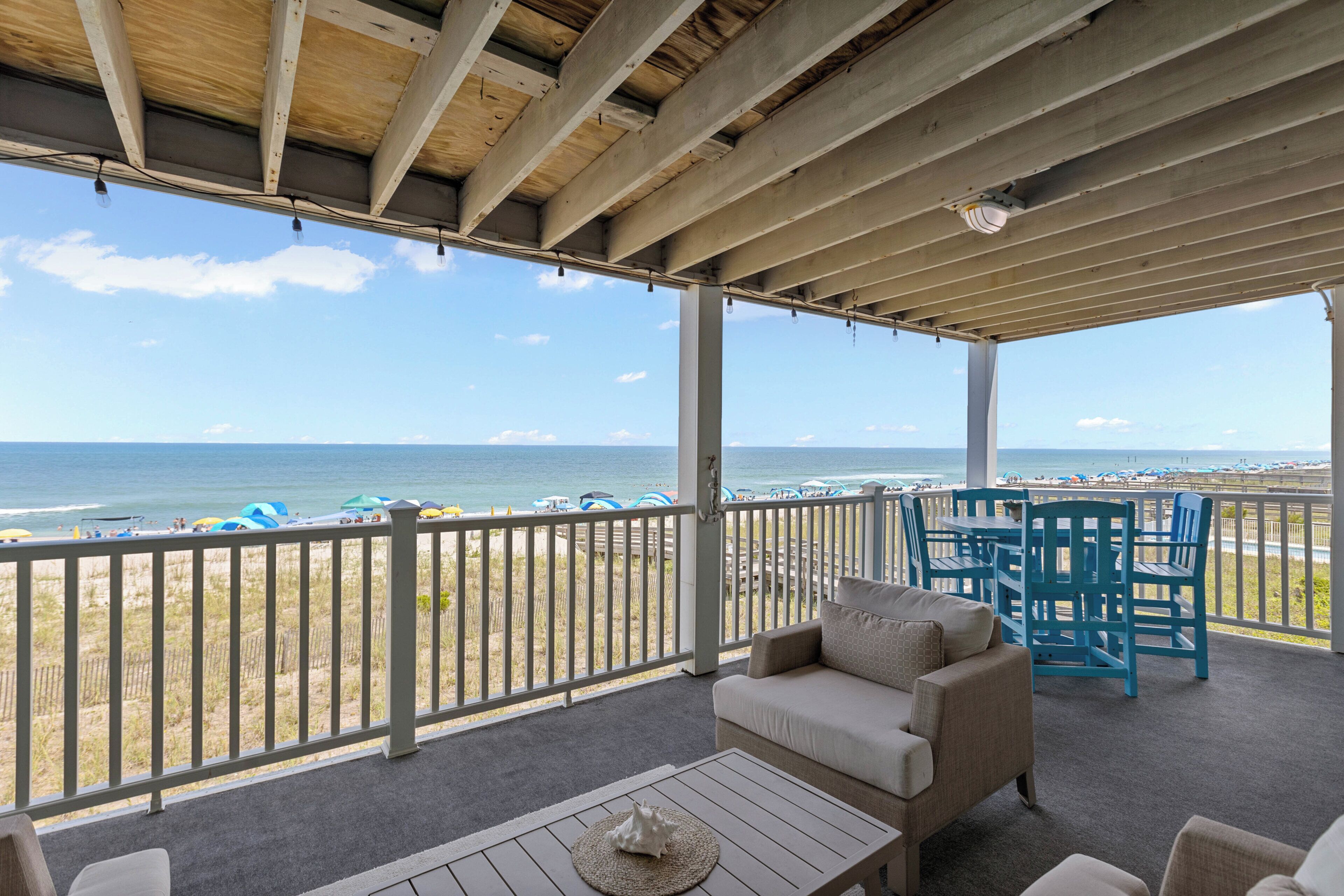 A covered outdoor area with a view of the beach, a sofa, chairs, and a table.