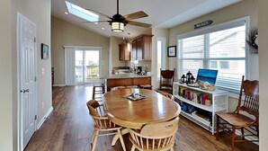 A spacious kitchen with wooden cabinets, a dining table with chairs, a ceiling fan, and a view of the beach through large windows. - Pelican Nest by Topsail Realty (Sneads Ferry)