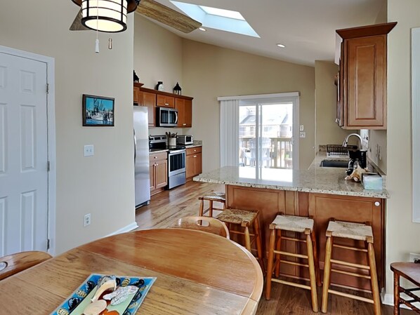 A modern kitchen with wooden cabinets, a large island with stools, and a skylight above the dining area. - Pelican Nest by Topsail Realty (Sneads Ferry)