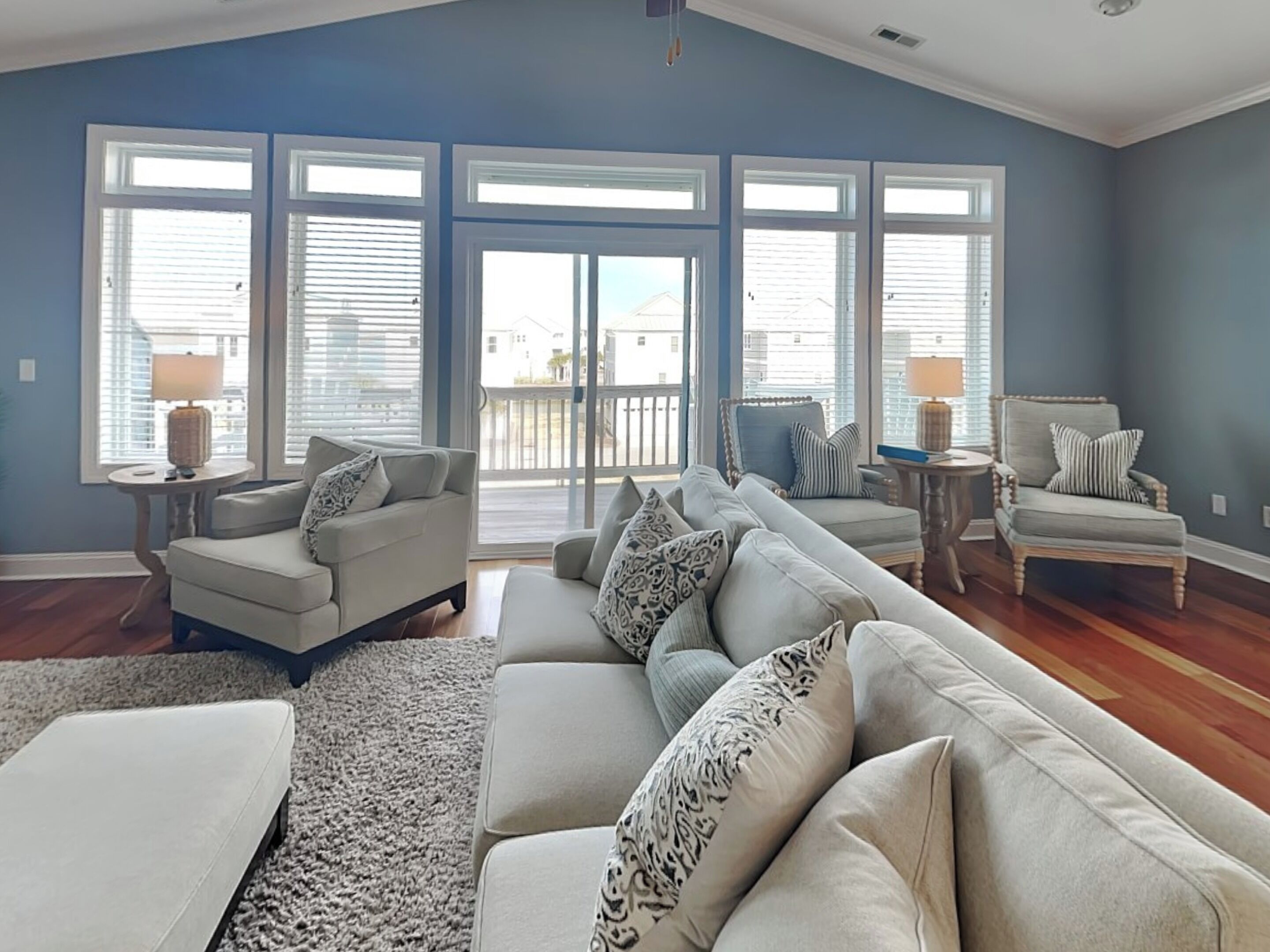 A spacious living room with a large sectional sofa, two armchairs, a coffee table, and a rug. There are large windows with blinds, a ceiling fan, and a view of a balcony.