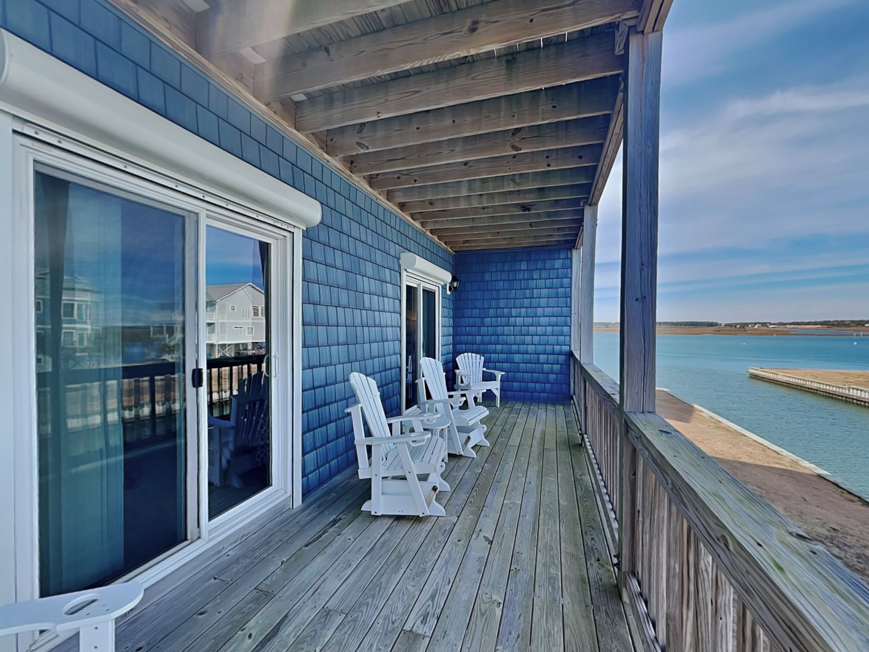 A covered deck with white Adirondack chairs, a wooden railing, and a view of the water.
