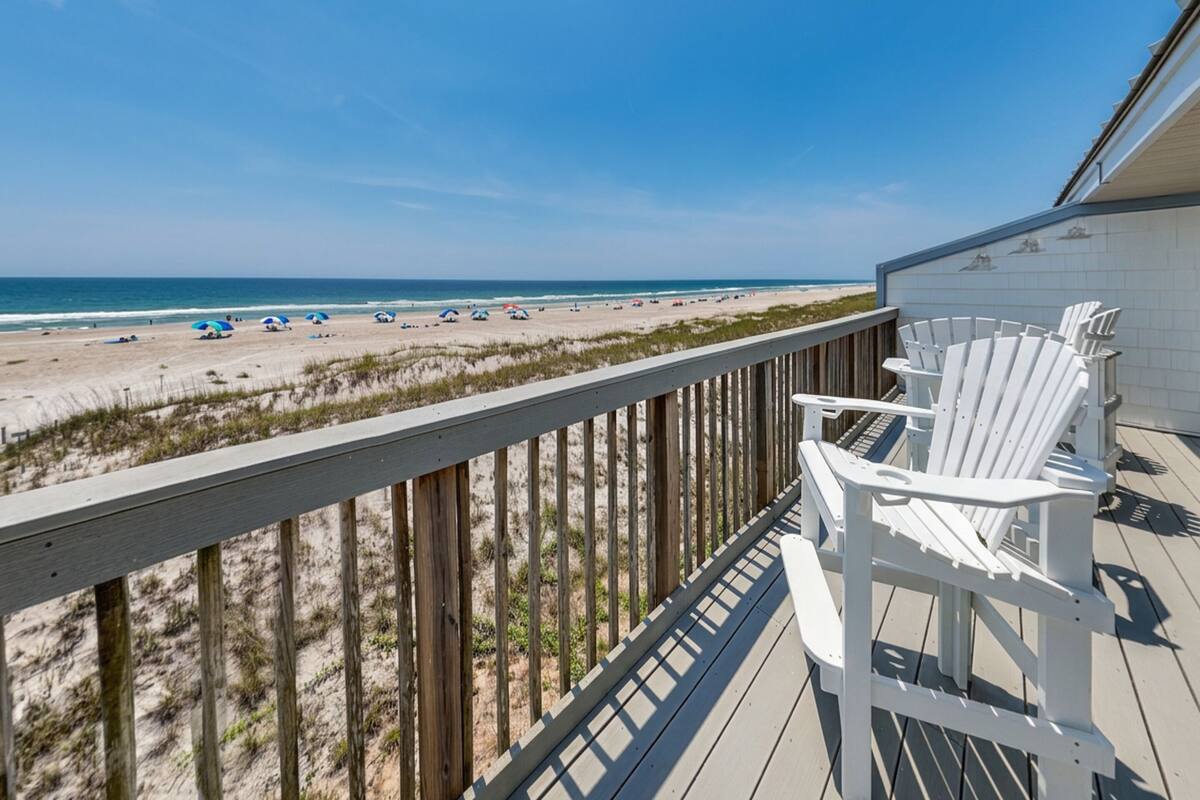 A wooden deck with white chairs overlooking a beach with umbrellas.