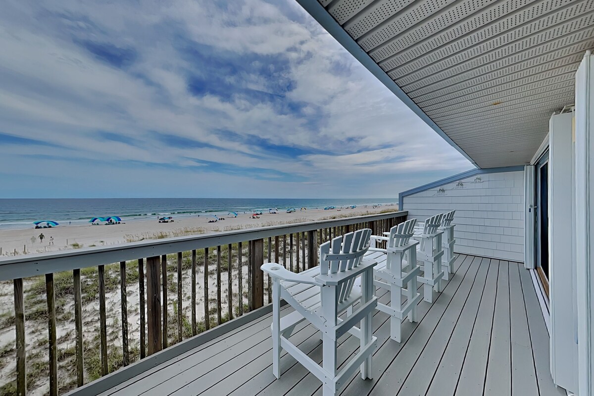 A wooden deck with white Adirondack chairs overlooking a beach with umbrellas and people.