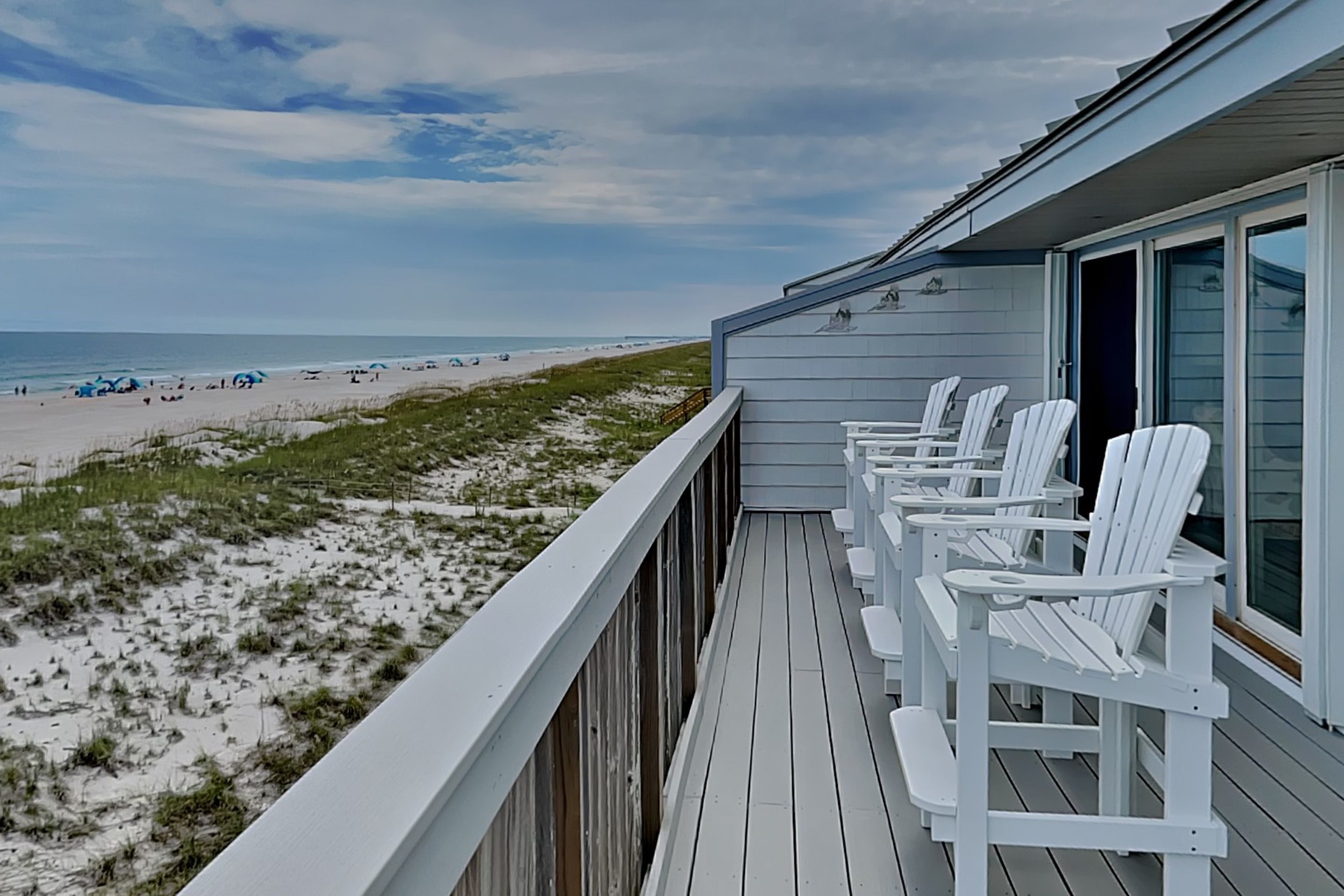 A deck with white chairs overlooking a beach with dunes and the ocean.