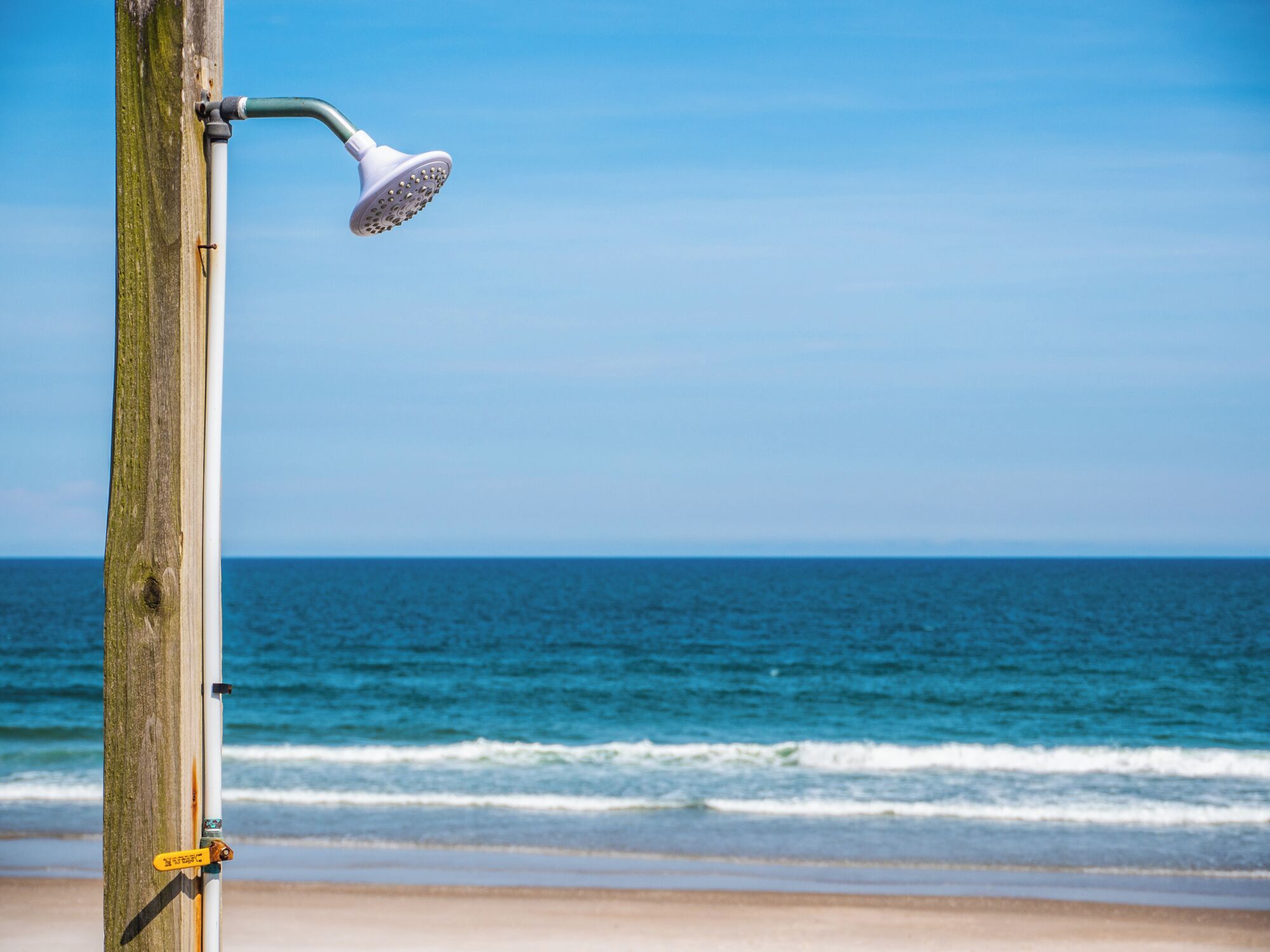 Ein Holzbalken mit daran befestigtem Duschkopf, mit Blick auf einen Strand, Wellen und einen klaren Himmel.
