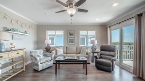 A spacious living room with a ceiling fan, a sofa, a chair, and a coffee table. There are two windows with blinds, a desk with a lamp, and a view of the outdoors through large sliding glass doors. - Coral Surf #A1 - Beachy Keen by Carolina Retreats (Carolina Beach)