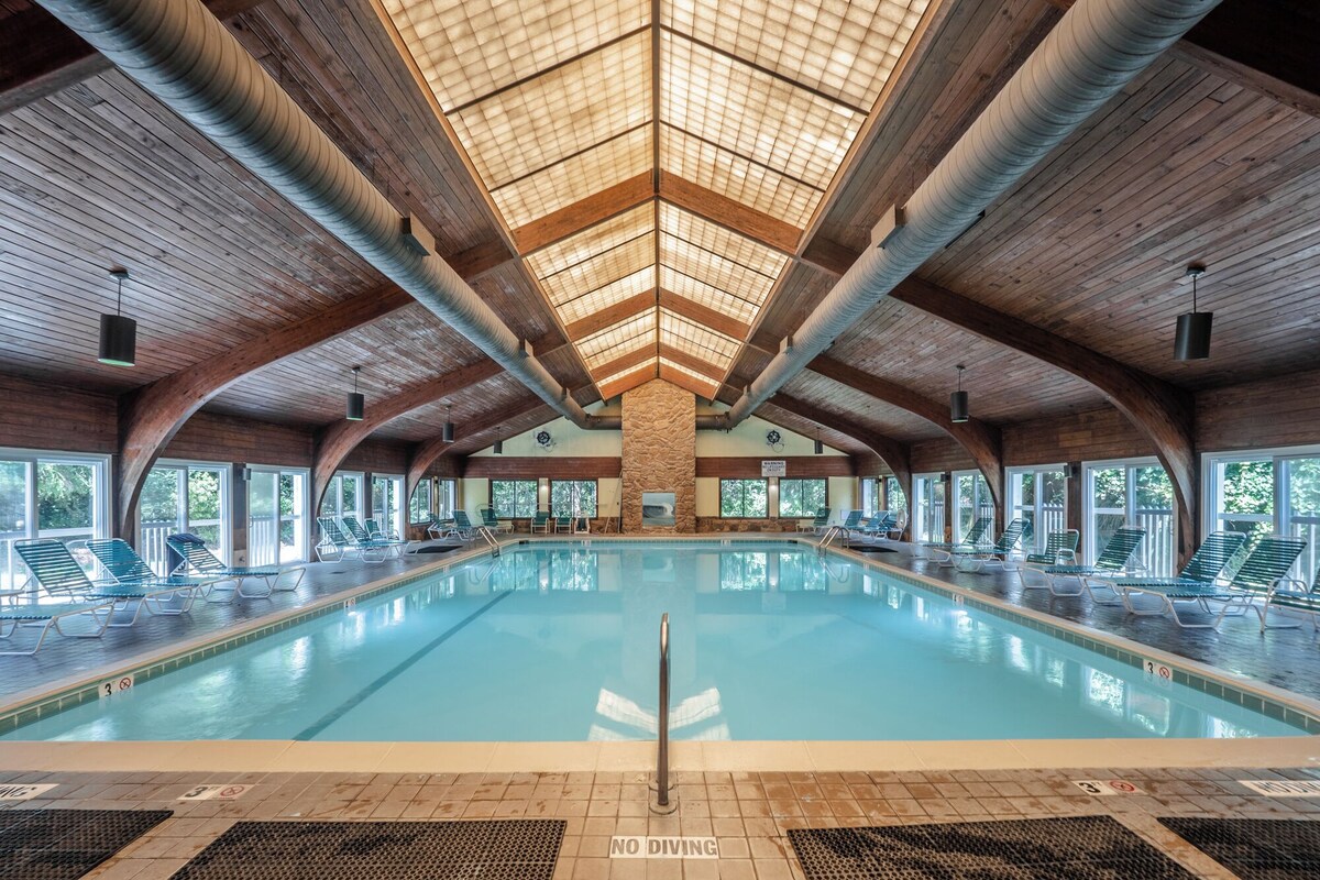 Une vaste piscine intérieure dotée d’un haut plafond et de poutres en bois, entourée de chaises longues et de grandes fenêtres.