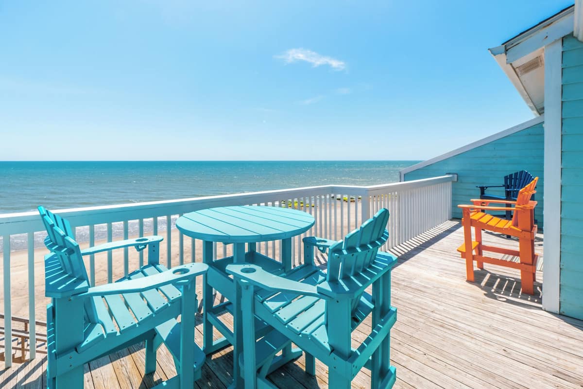 A wooden deck with turquoise chairs and a table, overlooking a beach.