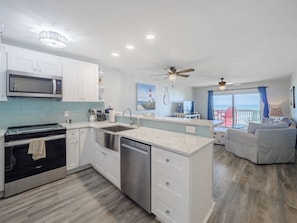 A modern kitchen with white cabinets, a stainless steel range, and a dishwasher. There is a dining area with a view of the ocean and a balcony with red chairs. - Ocean Dunes by Carolina Retreats (Kure Beach)