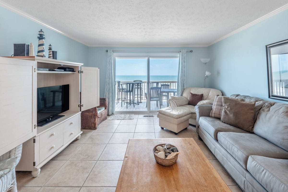 A living room with a TV, sofa, and a view of the sea through a glass door.