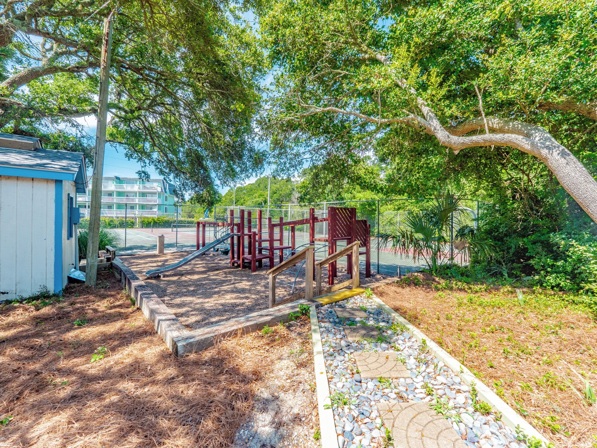 A playground with a slide and climbing structures, surrounded by trees and a small building.
