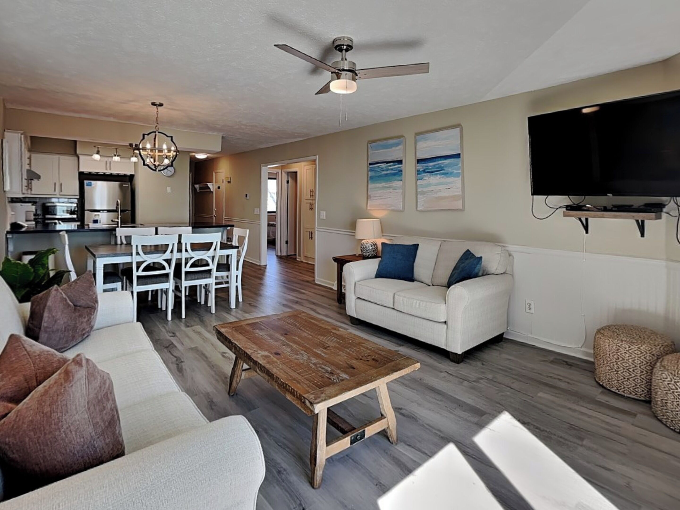 A spacious living room with a white sofa, a wooden coffee table, and a dining area with white chairs. A ceiling fan and a flat-screen TV are mounted on the wall.