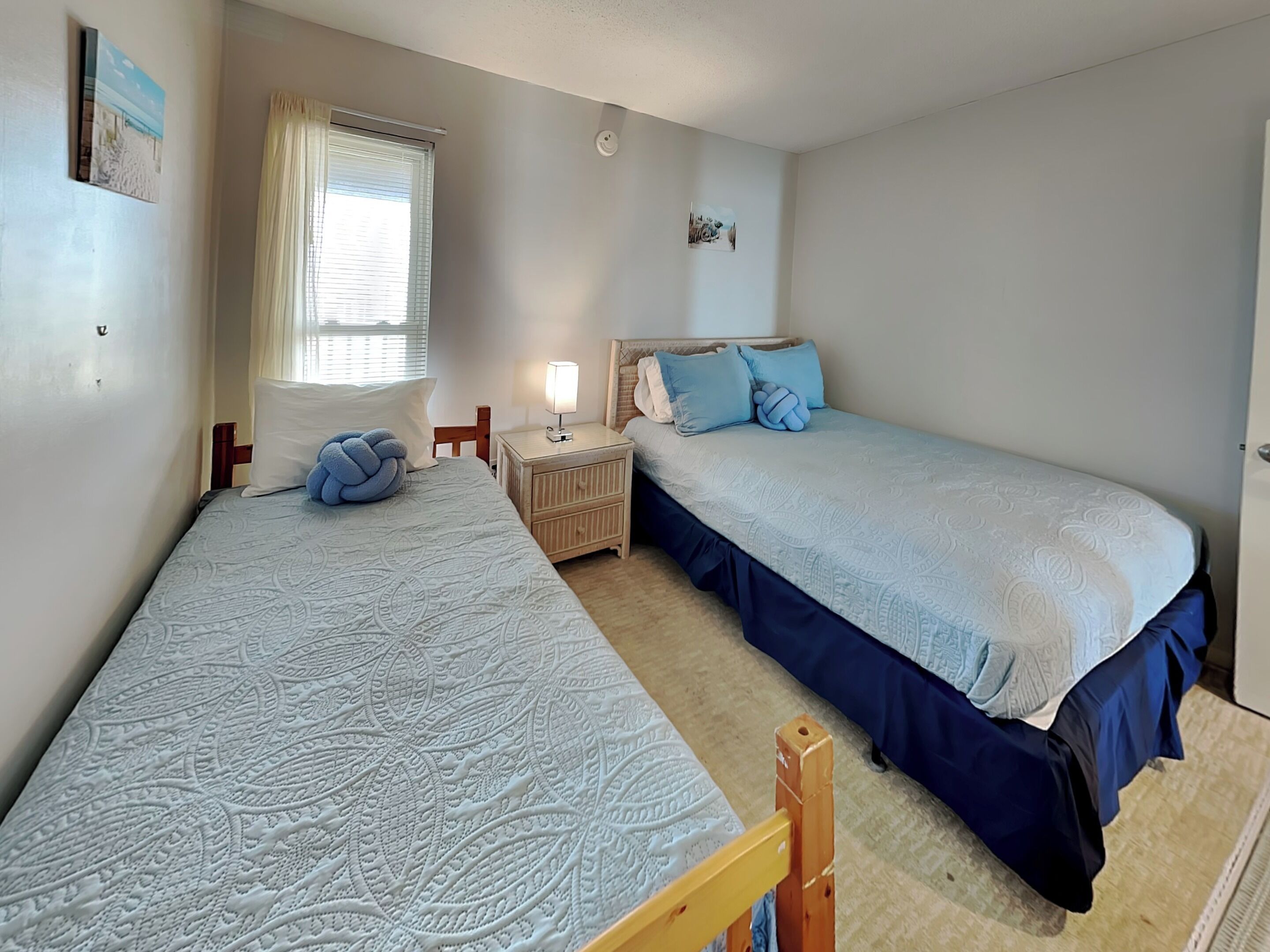 Ein Schlafzimmer mit zwei Einzelbetten, einem Nachttisch mit Lampe, einem Fenster mit Vorhängen und einem an der Wand montierten Rauchmelder.