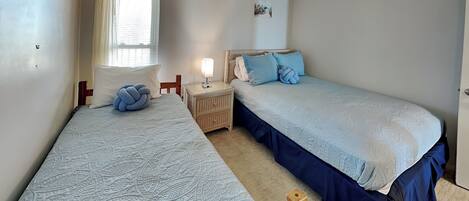 Ein Schlafzimmer mit zwei Einzelbetten, einem Nachttisch mit Lampe, einem Fenster mit Vorhängen und einem an der Wand montierten Rauchmelder.