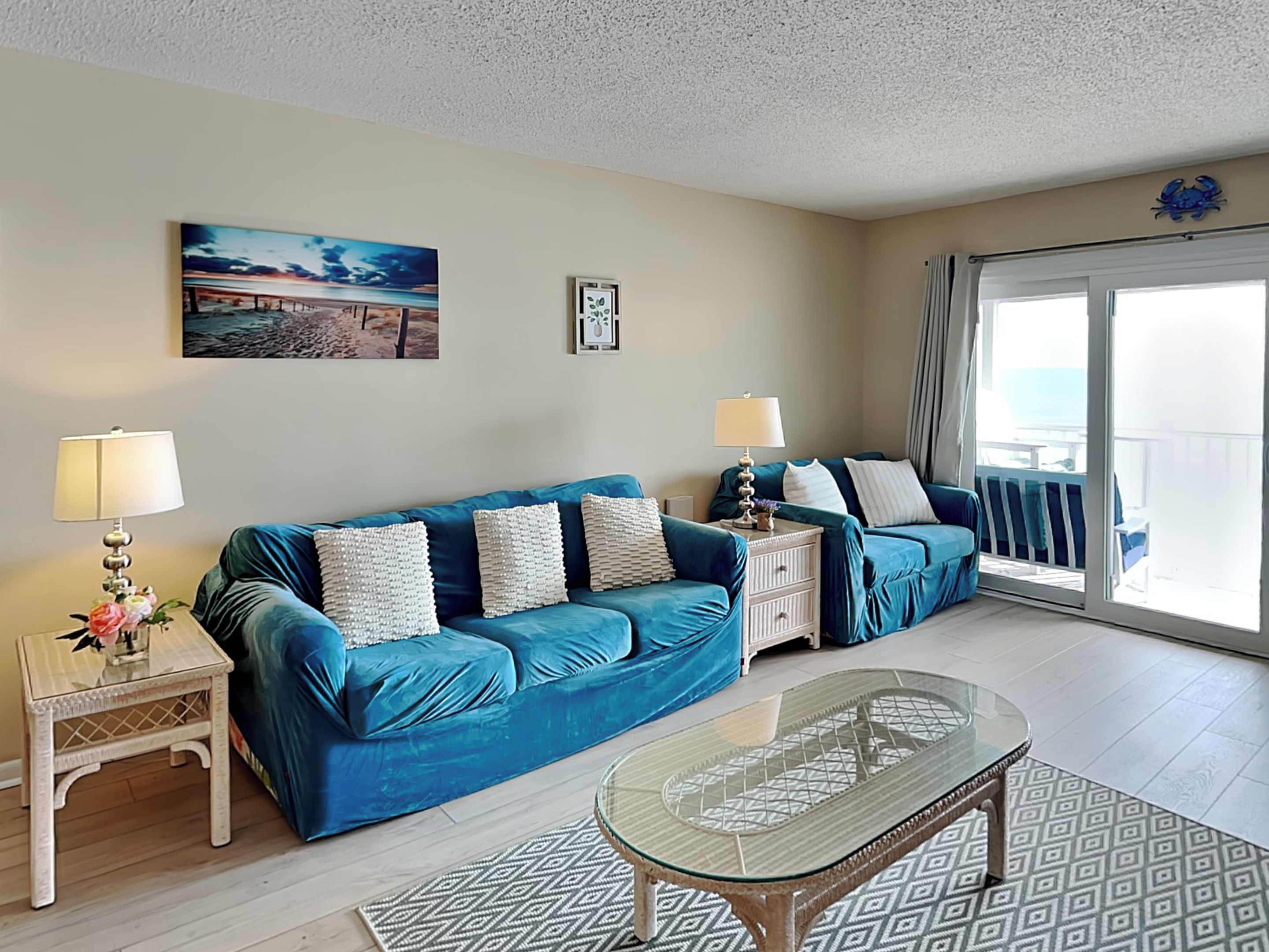 A living room with a blue sofa, two side tables with lamps, a glass coffee table, and a view of the sea through a sliding glass door.