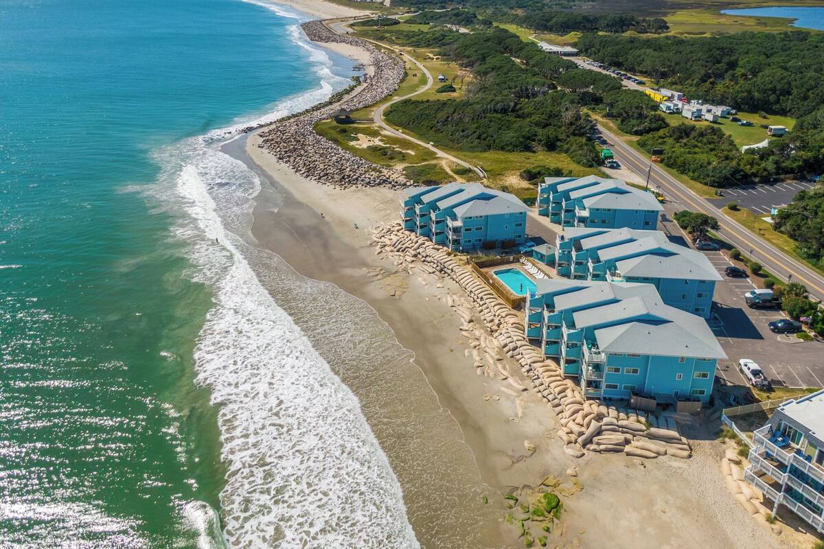 An aerial view of a coastal hotel with multiple blue buildings, a sandy beach, and a clear view of the ocean.
