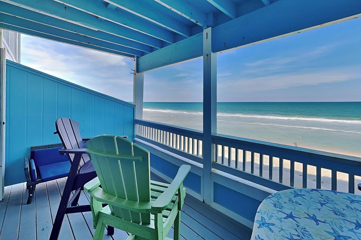 A covered deck with a view of the beach, two Adirondack chairs, and a table with a star pattern.