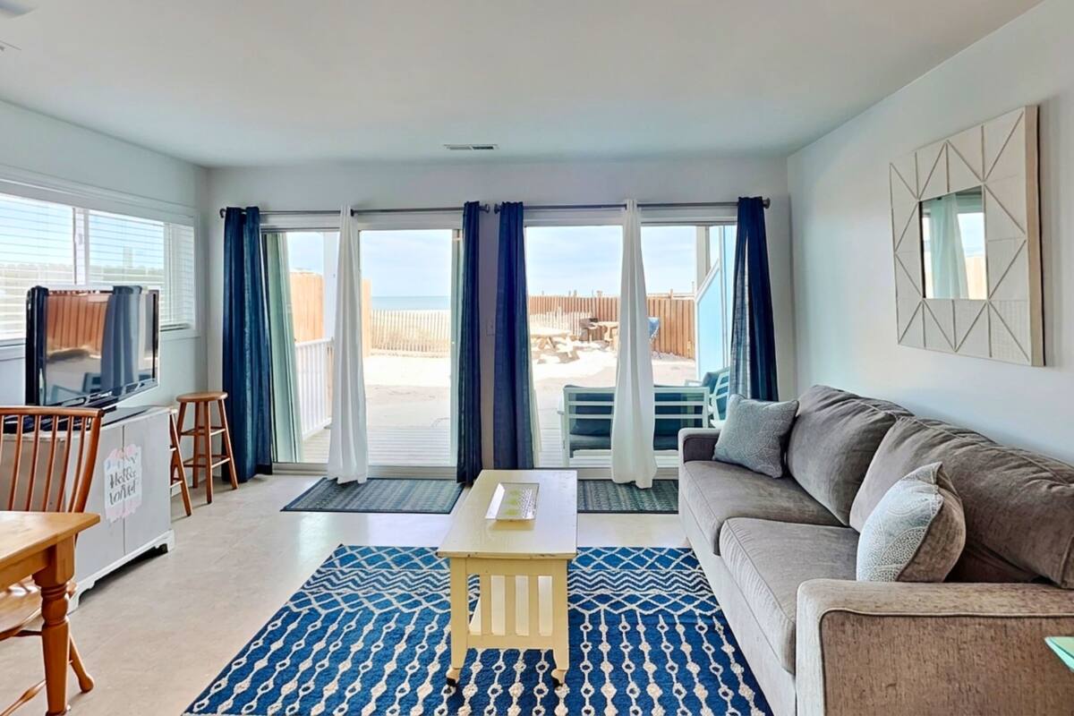 A living room with a sofa, a TV, a table, and a view of the beach through large sliding doors.