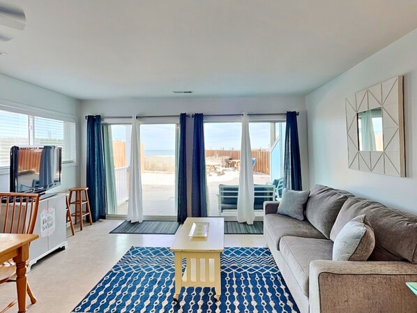 En stue med sofa, fjernsyn, bord og udsigt til stranden gennem store skydedøre.