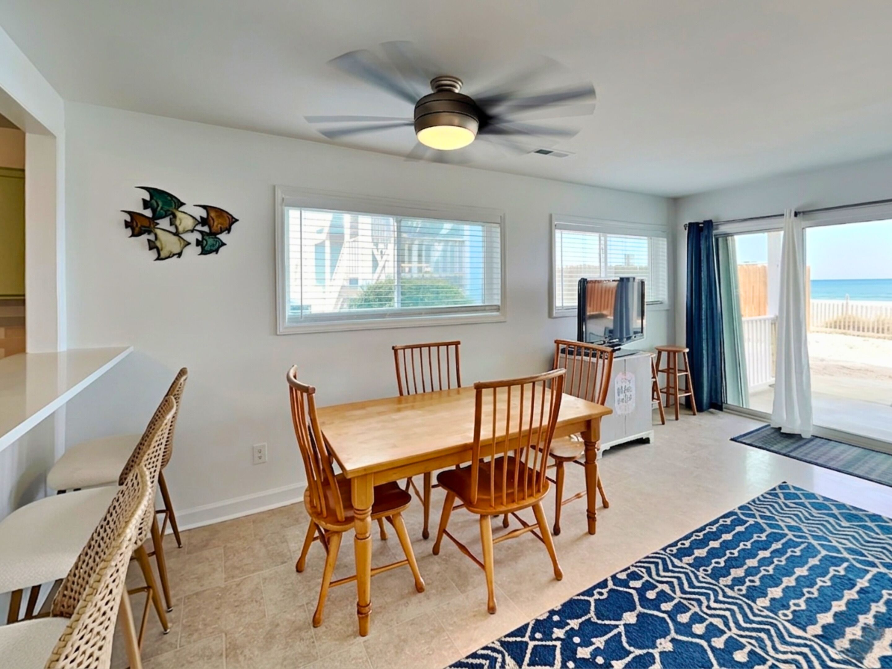 A dining area with a wooden table and chairs, a ceiling fan, and a view of the beach through large windows.