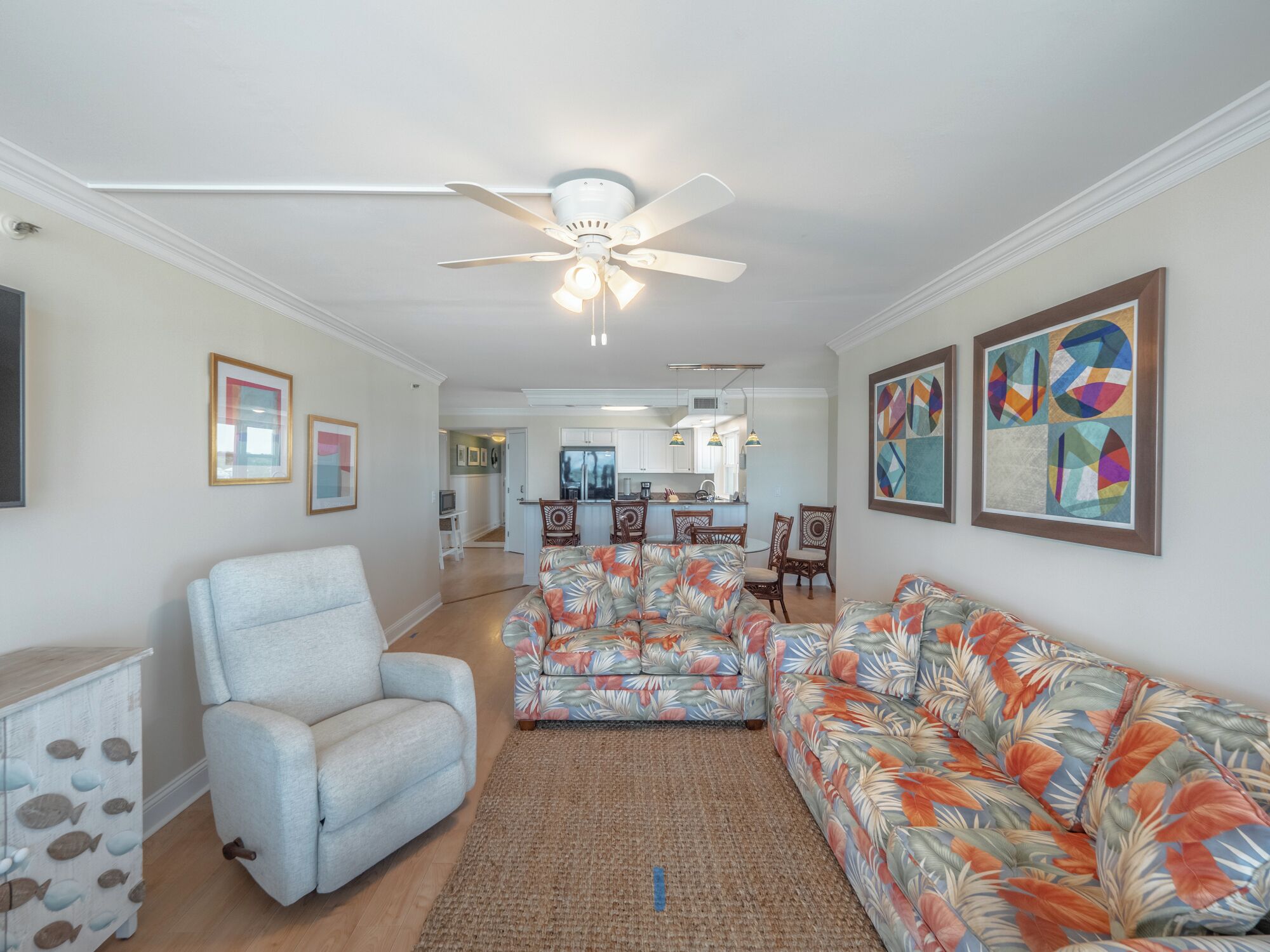 A living room with a ceiling fan, a sofa, a chair, and a rug. There is a kitchen in the background.