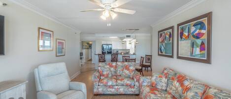 A living room with a ceiling fan, a sofa, a chair, and a rug. There is a kitchen in the background.